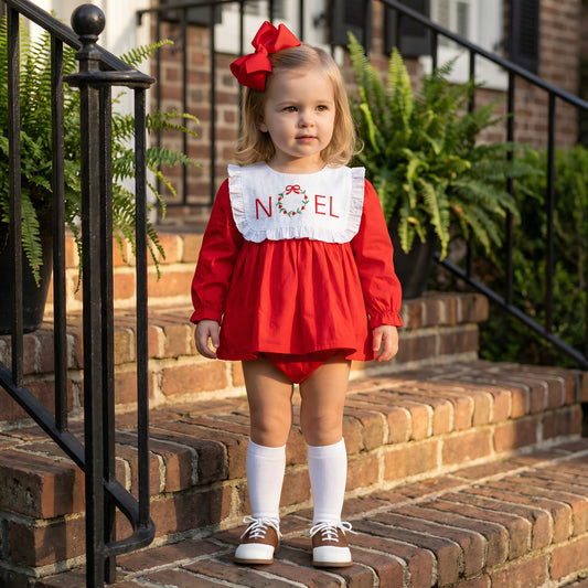 A full outdoor shot of a toddler girl with blonde hair and a large red bow, standing on brick steps. She wears a red long-sleeved romper with a white bib collar embroidered with "NOEL" and a wreath, white knee-high socks, and brown and white saddle shoes. Her hands are at her sides, and she looks slightly off-camera. The background shows a black metal railing, potted ferns, and the brick wall of a house.