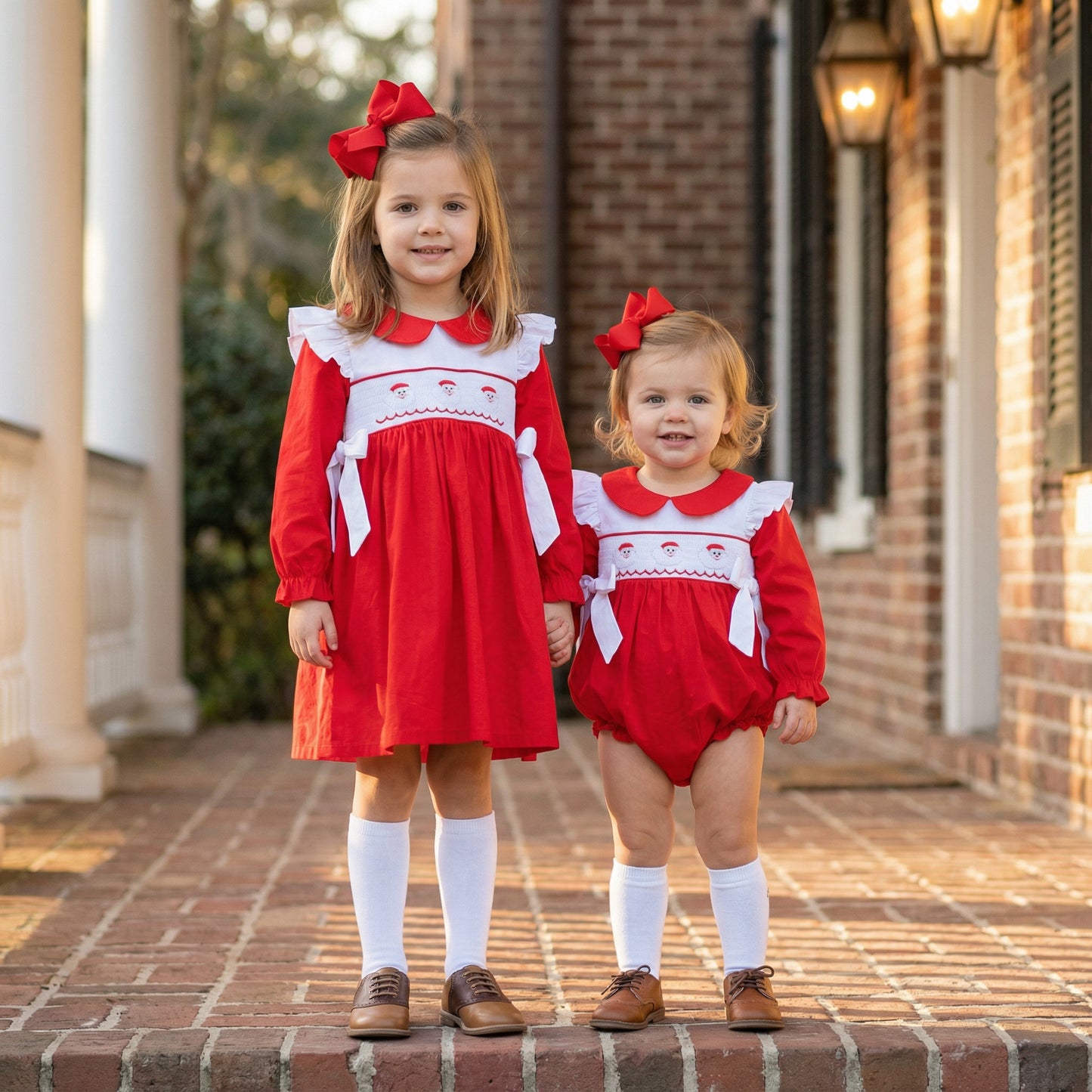 Two young girls standing hand-in-hand on a brick porch, wearing matching red holiday outfits with white smocked bodices featuring embroidered Santa faces. The older girl wears a dress, while the younger toddler wears a bubble romper; both have red hair bows, white knee socks, and brown shoes.