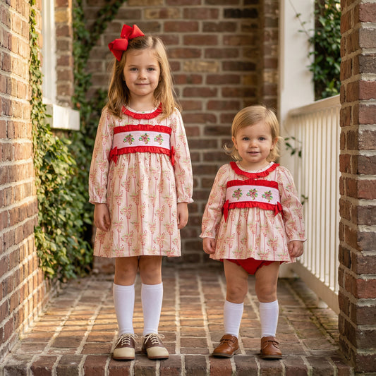 Two young girls, the older on the left and the younger on the right, pose side-by-side on a brick walkway. The older girl has long blonde hair with a large red bow, wearing a long-sleeved dress with pink floral patterns, red trim, smocking, white knee-high socks, and brown and beige saddle shoes. The younger girl has short blonde hair and wears a similar, shorter dress as a romper over red bloomers, with white knee-high socks and brown shoes.