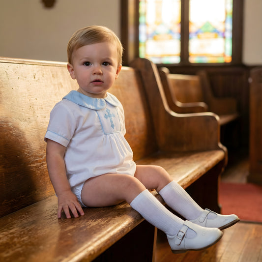 Toddler boy sitting on a wooden church pew wearing the Sunday Best - Hand Smocked Hand Embroidered Boys Blue Cross Bubble outfit with white t-strap shoes and stained glass in the background.