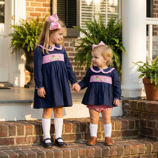 Two young sisters holding hands while standing on outdoor brick steps in front of a house. The older girl on the left wears a navy blue dress with a pink ruffled collar and a pink smocked chest panel embroidered with a nativity scene, paired with a large pink hair bow, white knee socks, and black Mary Jane shoes. She looks down at the younger toddler on the right, who wears a matching navy and pink smocked outfit with pink bloomers, white socks, and brown shoes.