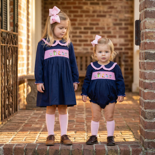 Two young girls stand side-by-side on a brick porch, wearing matching navy blue outfits with pink accents. The older girl on the left wears a long-sleeved dress with a pink ruffled collar and a pink smocked panel across the chest embroidered with a Nativity scene featuring Mary, Joseph, Jesus, and sheep. She wears pink knee-high socks and brown saddle shoes. The younger toddler on the right wears a matching navy blue bubble romper with the same pink collar and Nativity smocking.