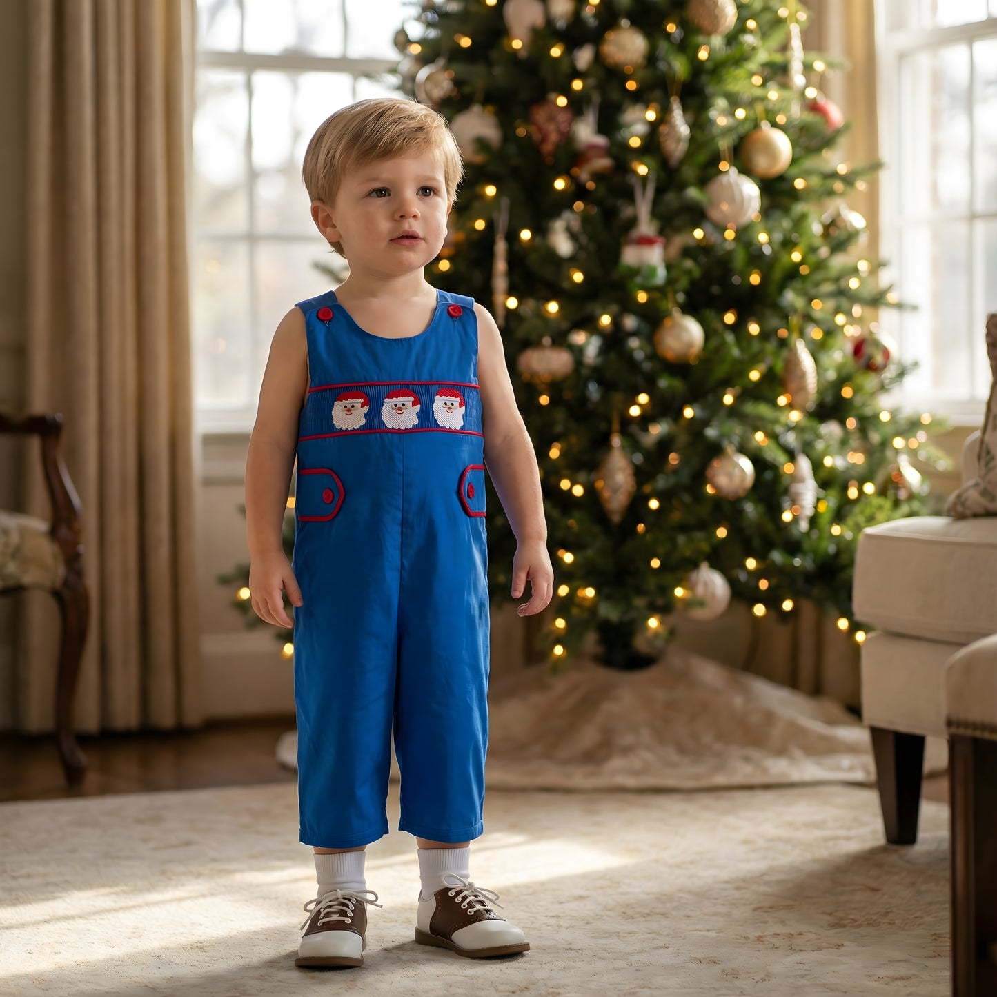 Toddler boy wearing a Boys Navy Blue Santa Smocked Christmas Jon Jon with saddle shoes standing in front of a decorated tree.