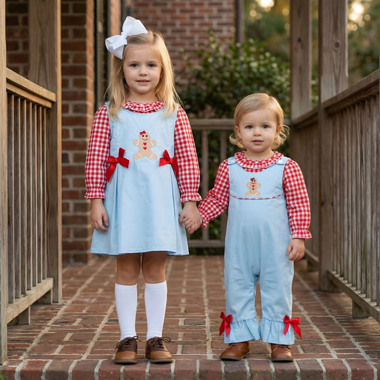 Two toddler girls wearing matching light blue and red gingham holiday outfits with gingerbread man appliqués, one in a dress and one in a romper, holding hands on a brick porch.