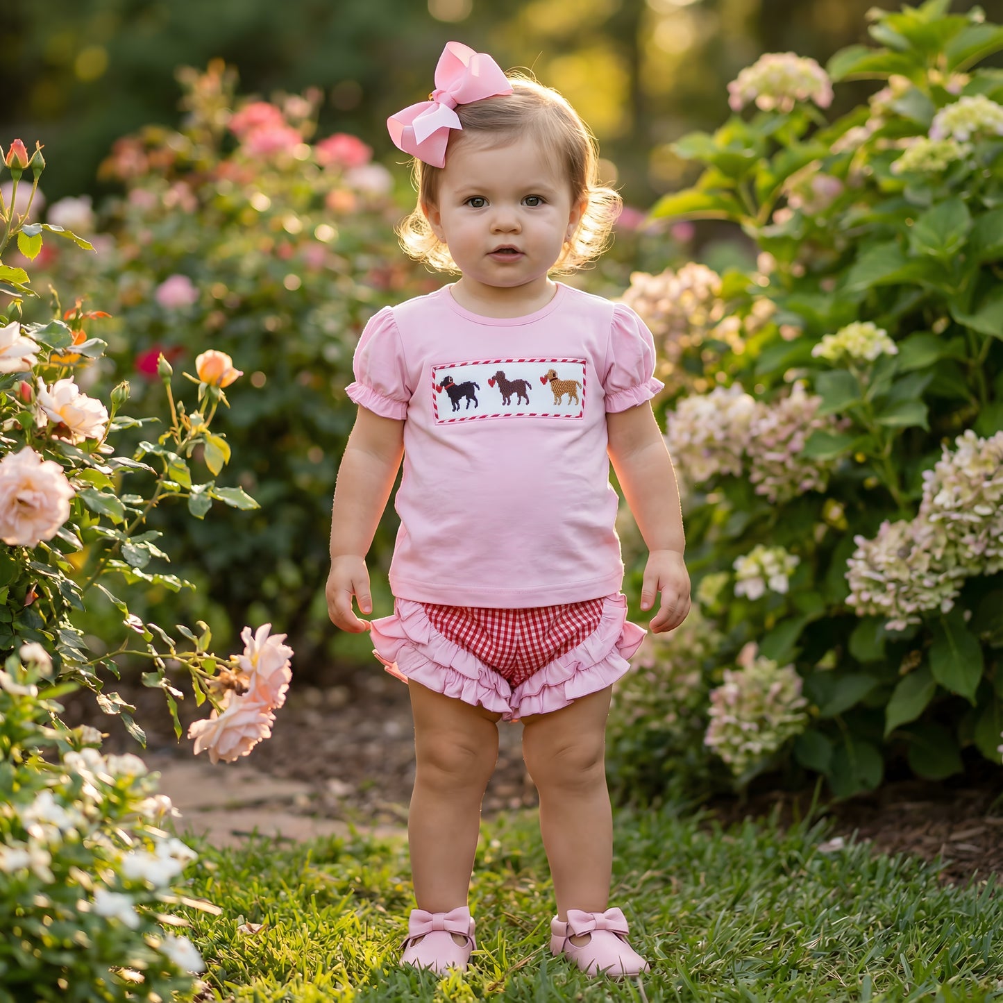 Toddler girl modeling the Hand Smocked Valentines Puppy Dogs - Hand Smocked Girls Set, featuring a pink top with embroidered puppies holding red hearts and red gingham ruffle bloomers.