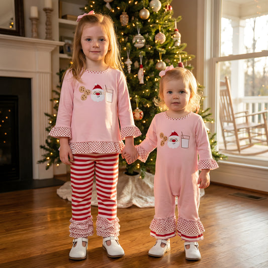 Two sisters holding hands in front of a Christmas tree wearing matching pink Girls Milk And Cookies With Santa Christmas Embroidered Pants Set and Romper.