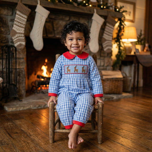 Toddler boy wearing blue gingham hand smocked reindeer romper sitting by a Christmas fireplace.