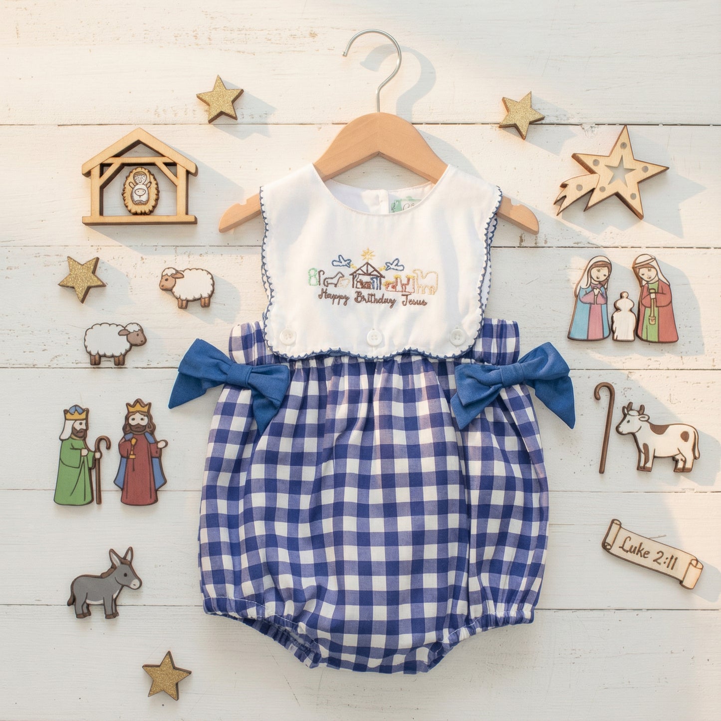 An overhead photograph captures a baby's romper on a wooden hanger, adorned with a "Happy Birthday Jesus" embroidered nativity scene on the white bodice and blue gingham shorts with blue bows. The romper is surrounded by various small wooden nativity figurines, including the holy family, shepherds, kings, animals, a stable, and stars, all arranged on a white wooden plank surface. A wooden scroll with "Luke 2:11" is also present.