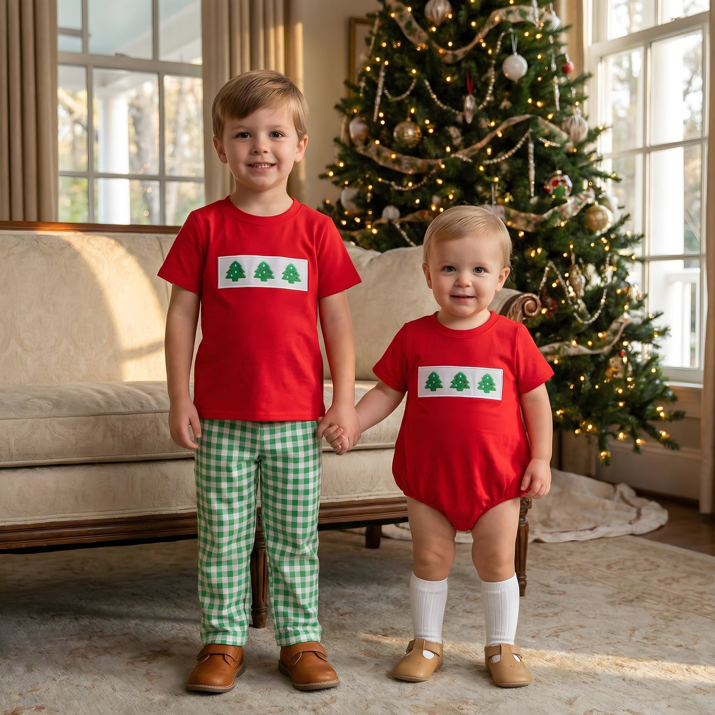 Two smiling brothers holding hands in front of a Christmas tree wearing matching red outfits with the Embroidered Christmas Tree Trio Boys design, featuring green gingham pants and a bubble romper.