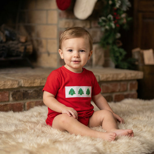 Baby boy wearing Smocked Peacock red Embroidered Christmas Tree Trio Boys Bubble romper by a fireplace.