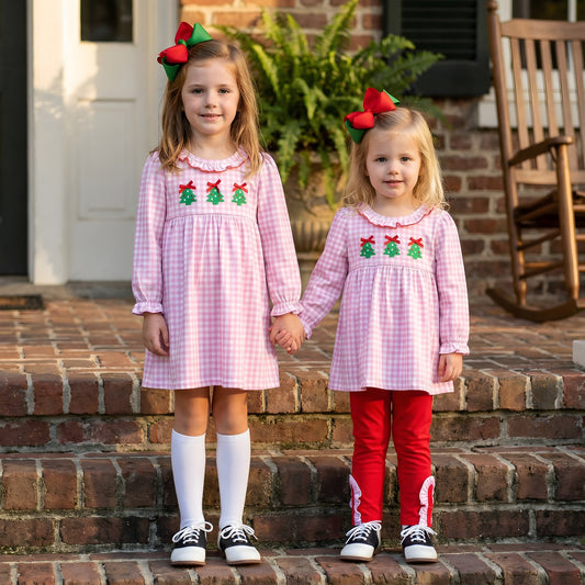 Two sisters in matching pink gingham smocked Christmas tree dresses holding hands on brick steps.