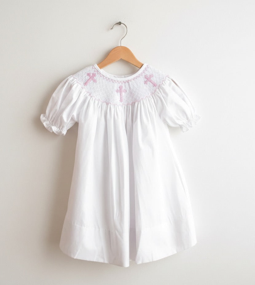 White girls smocked christening dress with pink crosses hanging on a wooden hanger against a white wall.
