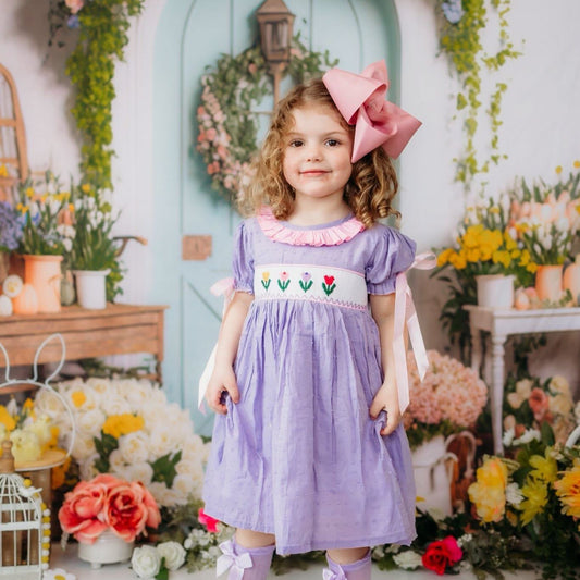A young girl stands in a whimsical floral room wearing a lavender puffy dress with a white embroidered bodice, a large pink bow in her hair, and matching lavender socks.