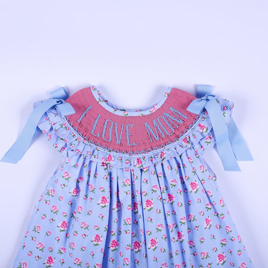 Close-up of pink hand-smocked yoke with blue I Love Mom embroidery, flutter sleeves, and shoulder ribbons on dress.