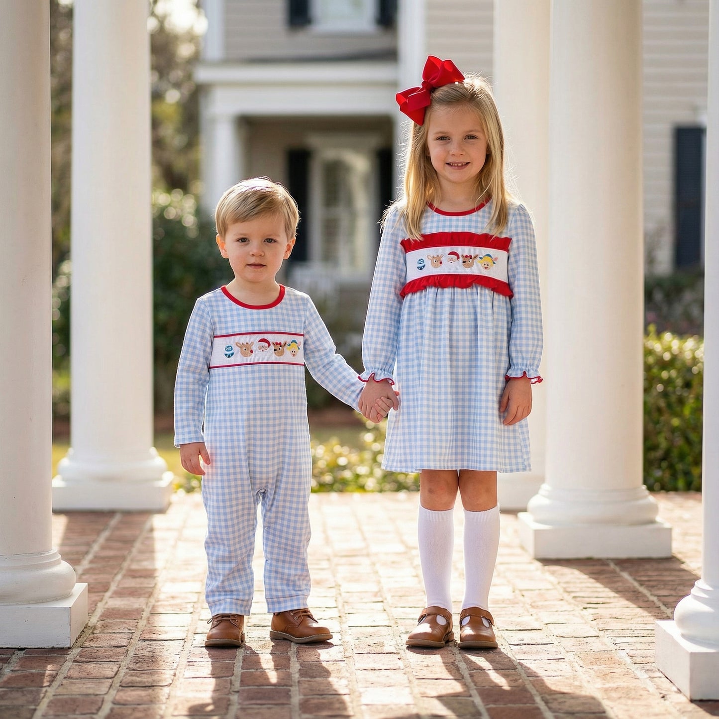 Siblings holding hands on porch in matching blue gingham smocked Christmas outfits with Rudolph, elf, and snowman.