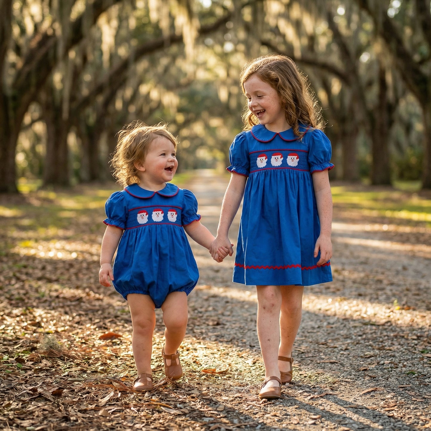 Two sisters walking holding hands outdoors wearing matching navy blue Santa smocked dress and romper for Christmas.