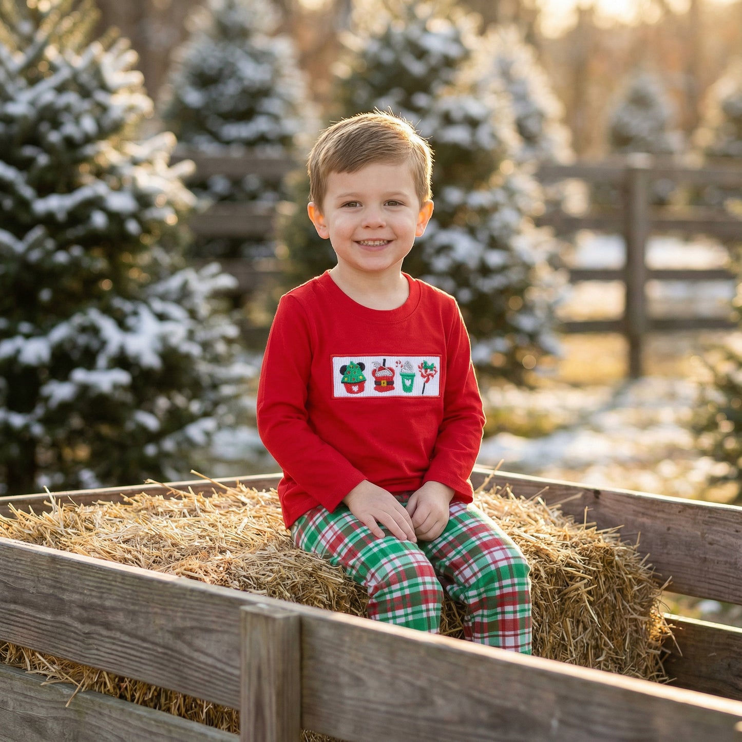 Boy sitting on hay wagon at tree farm wearing red smocked Christmas treats top and plaid pants.