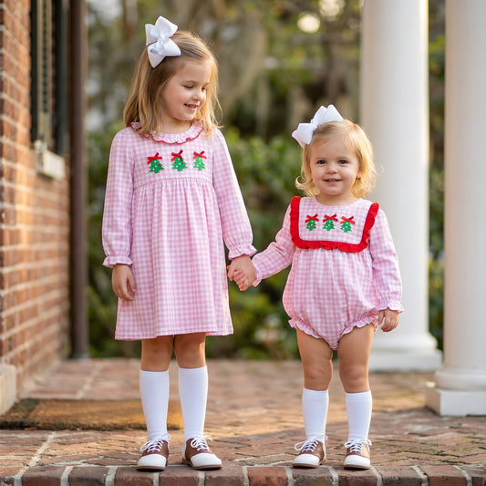 Two young girls, sisters, hold hands in matching pink gingham smocked Christmas tree outfits. One in a dress, the other a romper.