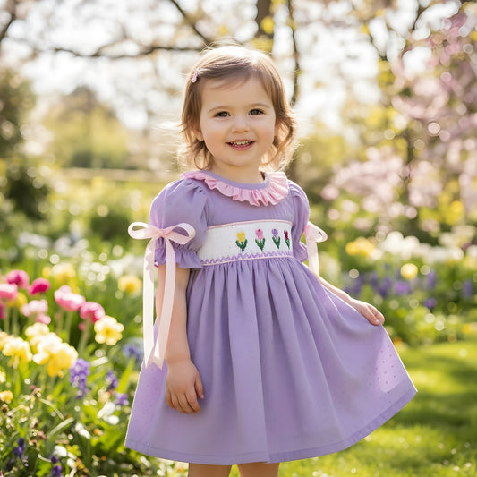 Little girl in a lavender smocked Easter dress with embroidered tulips and pink collar standing in a spring garden.