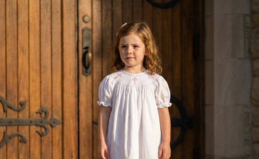 Young girl wearing a white smocked christening dress with pink crosses, standing by wooden church doors.