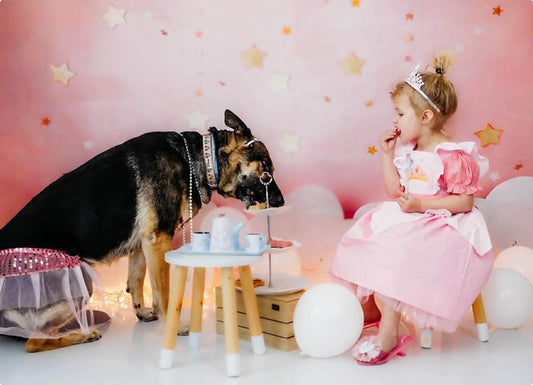 Young girl in a pink princess dress and a German Shepherd wearing a pink tutu having a tea party together against a starry pink background.