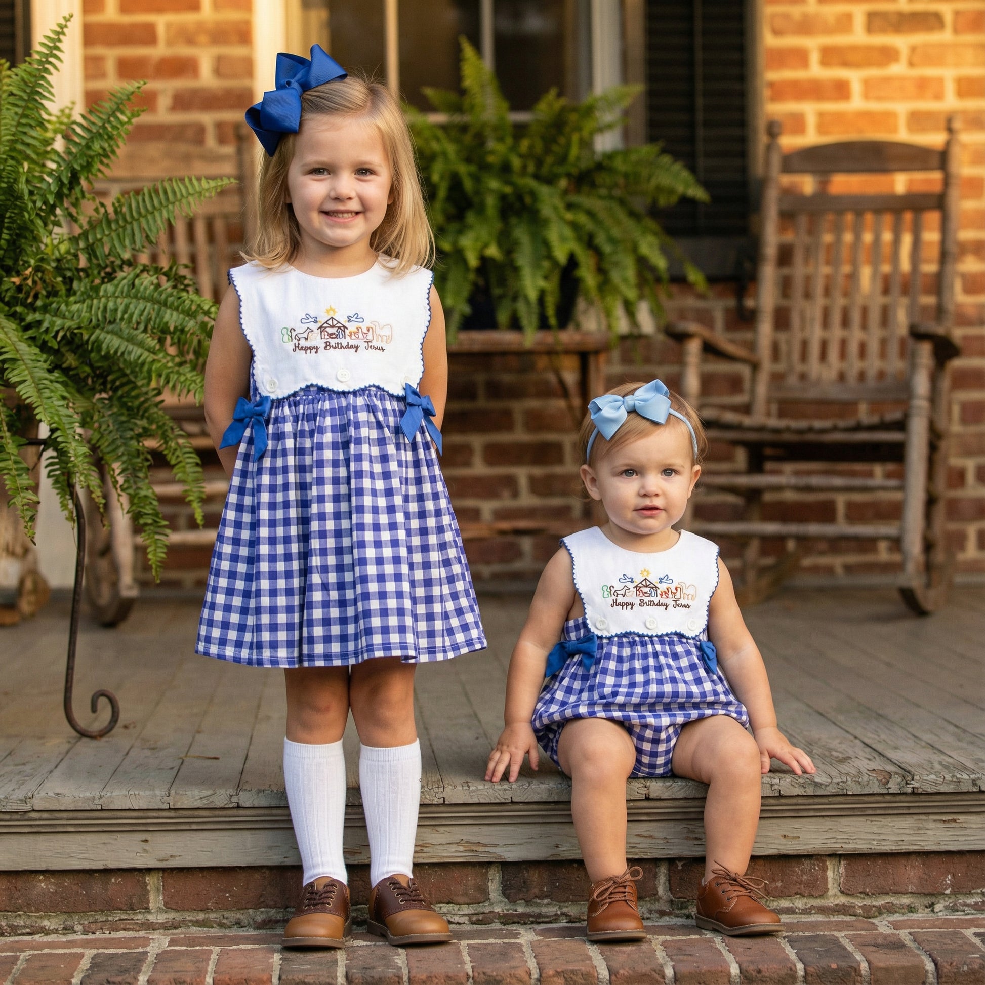 A photograph of two young girls, one standing and one sitting on a rustic wooden porch in front of a brick house. Both girls are wearing matching blue and white gingham outfits with embroidered white bibs that read "Happy Birthday Jesus" and blue hair bows. The older girl wears a dress with white knee-high socks and brown shoes, while the younger girl wears a romper and brown shoes. Ferns in pots and wooden rocking chairs are visible in the background.