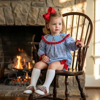 A toddler girl wearing a blue gingham romper with reindeer embroidery, a red hair bow, white knee-high socks, and saddle shoes sits in a wooden chair in front of a stone fireplace with a fire burning.