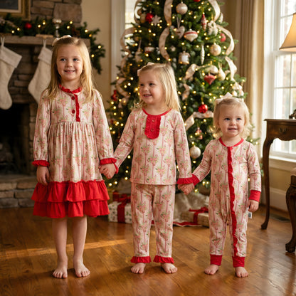 Three toddler girls pose together in matching soft bamboo Christmas sleepwear, featuring a nightgown, two-piece pajama set, and zippy with red accents, in front of a decorated Christmas tree and fireplace.