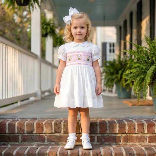 A toddler girl wears a white smocked dress with pink embroidered floral baskets, a large white hair bow, white ruffle socks, and white t-strap shoes, standing on brick porch steps. The traditional boutique-style dress is perfect for spring and summer fashion.