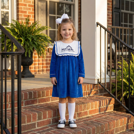 A young girl with blonde hair smiles while standing on brick steps, wearing a long-sleeved royal blue velvet dress with a large white ruffled collar embroidered with a navy blue nativity scene. She is accessorized with a large white hair bow, white knee-high socks, and navy and white saddle shoes.
