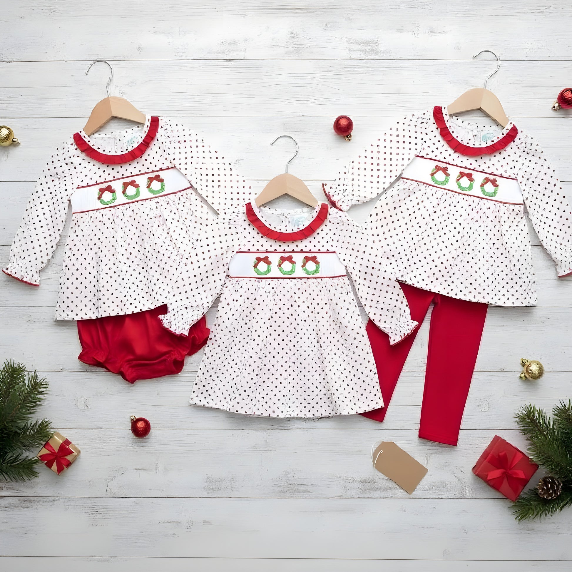 Three children's outfits with a white fabric, red dot pattern, and embroidered Christmas wreaths on the smocking are laid out on wooden hangers against a white wooden plank background. The left outfit is a dress and red bloomers, the middle is a dress, and the right is a dress with red leggings. Christmas decorations are scattered around the outfits.