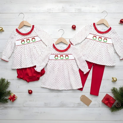 Three children's outfits with a white fabric, red dot pattern, and embroidered Christmas wreaths on the smocking are laid out on wooden hangers against a white wooden plank background. The left outfit is a dress and red bloomers, the middle is a dress, and the right is a dress with red leggings. Christmas decorations are scattered around the outfits.