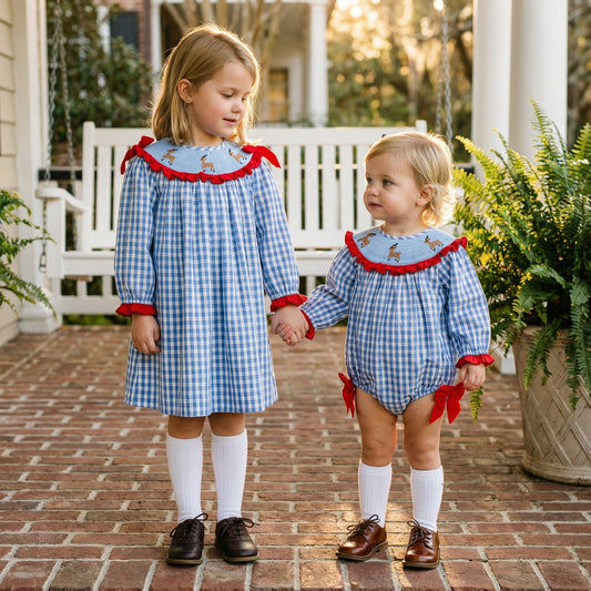 Two young children hold hands on a brick porch while wearing matching blue and white gingham outfits featuring red trim and reindeer embroidery.