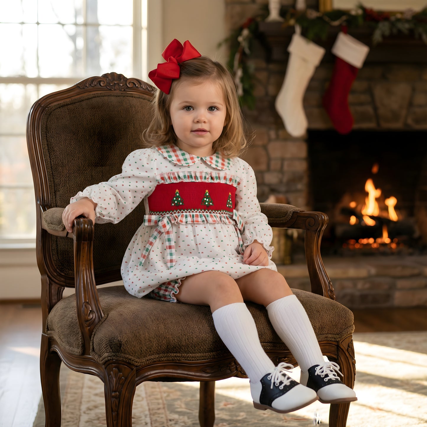 A young girl with light brown hair sits on an ornate wooden armchair, wearing a festive white dress with red smocking featuring embroidered Christmas trees and a plaid collar. She wears a large red bow in her hair, white knee-high socks, and black and white saddle shoes. The background features a cozy stone fireplace with a fire burning and Christmas stockings hanging from the mantle.