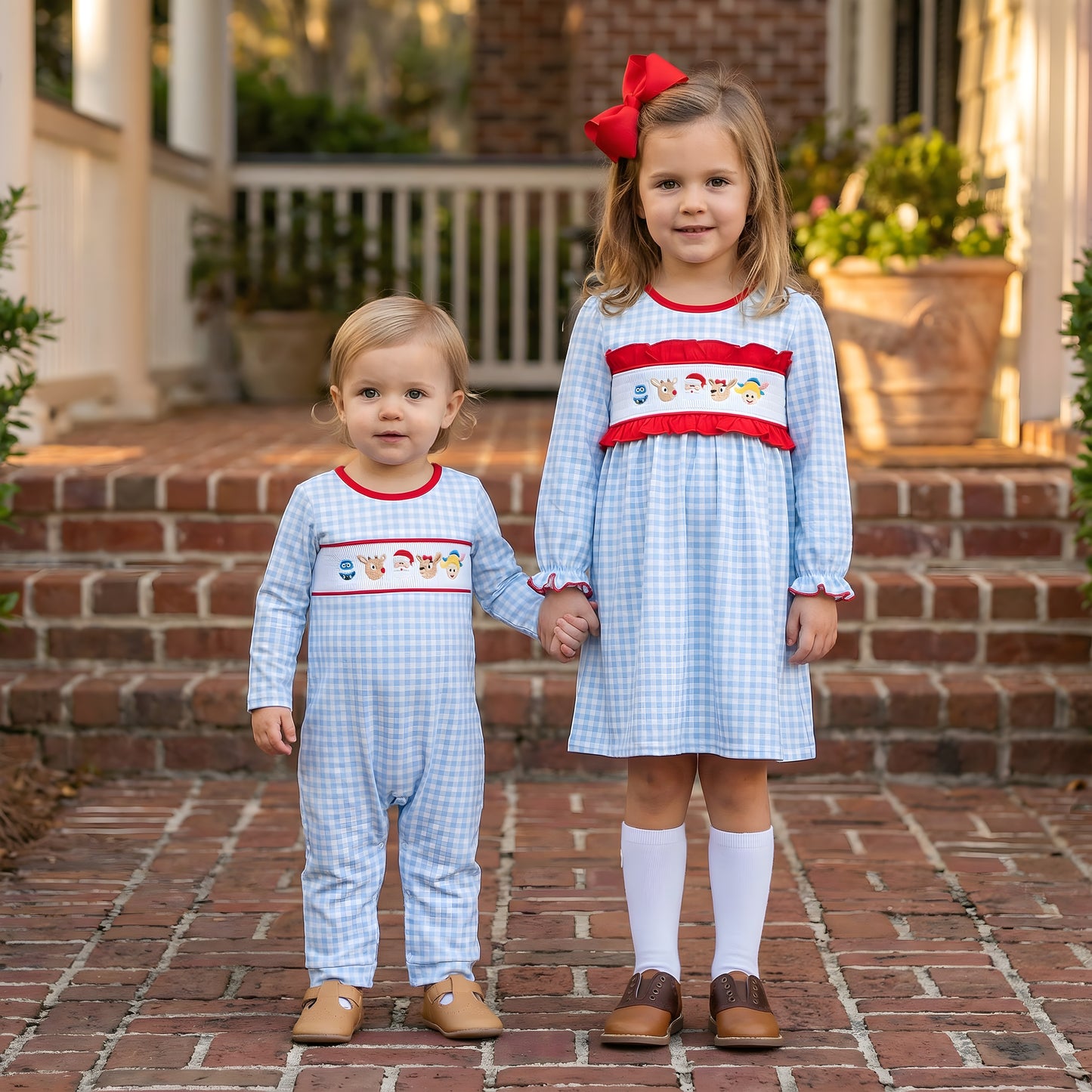 A toddler in a blue gingham smocked longall and a young girl in a matching smocked dress with Christmas motifs hold hands on brick steps. Both outfits feature red trim and embroidered Santa, reindeer, and snowman figures. The older girl wears a large red hair bow, white knee-high socks, and brown saddle shoes, while the toddler wears tan leather t-strap shoes. The setting is outdoors on a sunny day in front of a house with a white porch railing.