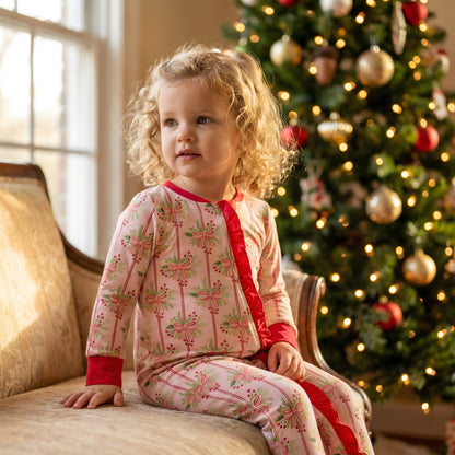Toddler girl wearing a pink bamboo Christmas zippy with bow print and red ruffles sitting in front of a lit holiday tree.
