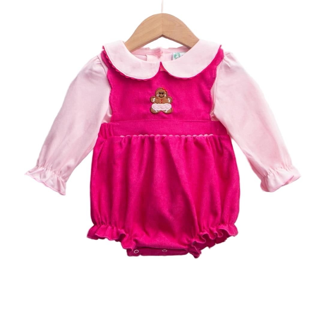A photograph of a baby outfit hanging on a wooden hanger against a white background. The outfit consists of a light pink long-sleeved shirt with a rounded white collar and ruffled cuffs, worn underneath a bright pink corduroy bubble jumper with a gathered waist, ruffled leg openings, and an embroidered brown gingerbread man figure in the center of the chest.