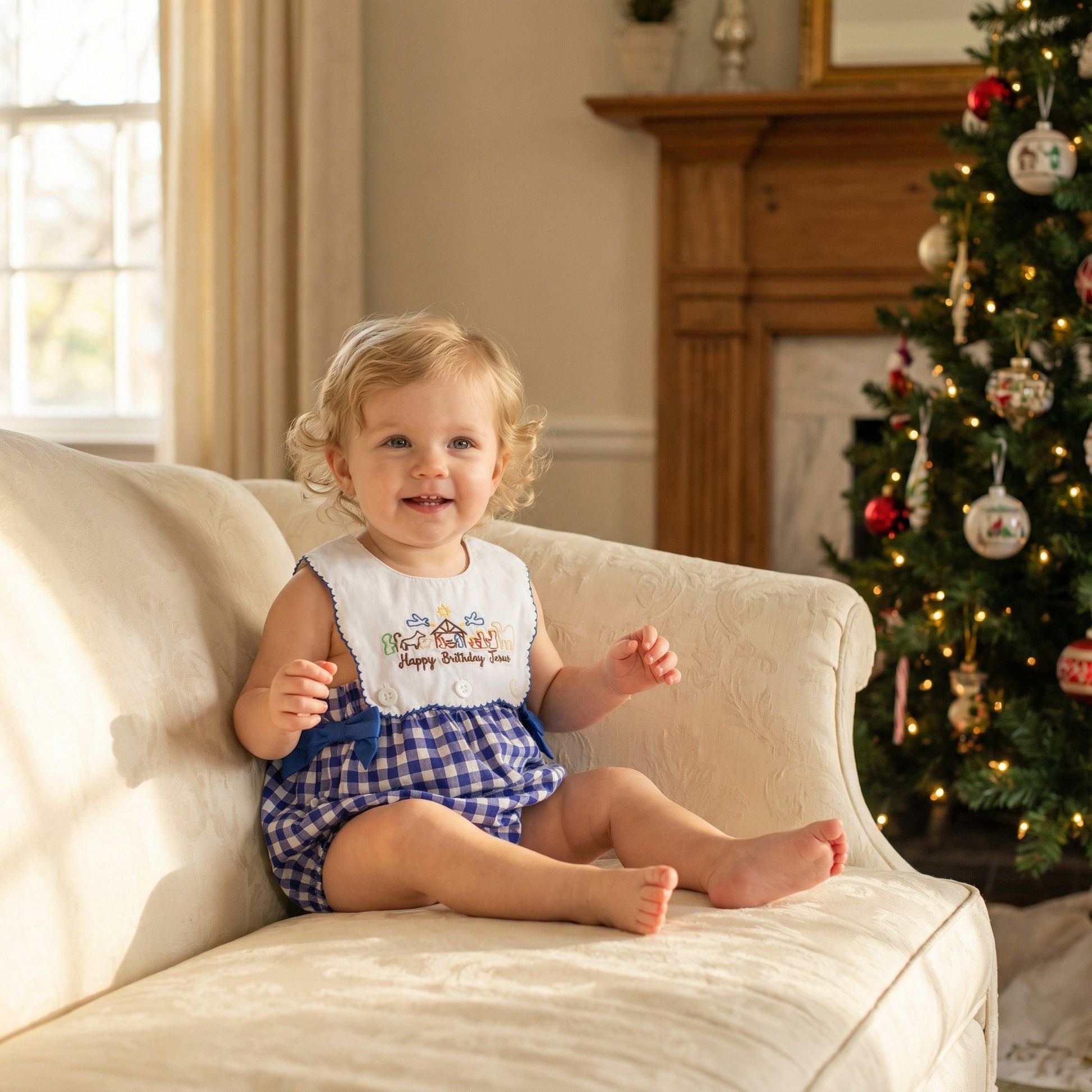 A young toddler with curly blonde hair and a big smile sits barefoot on a cream-colored sofa, wearing a blue and white gingham romper with an embroidered nativity scene bib that reads "Happy Birthday Jesus." In the background is a decorated Christmas tree and a wooden fireplace mantel, with sunlight streaming in from a window to the left.