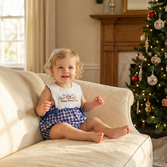 A young toddler with curly blonde hair and a big smile sits barefoot on a cream-colored sofa, wearing a blue and white gingham romper with an embroidered nativity scene bib that reads "Happy Birthday Jesus." In the background is a decorated Christmas tree and a wooden fireplace mantel, with sunlight streaming in from a window to the left.