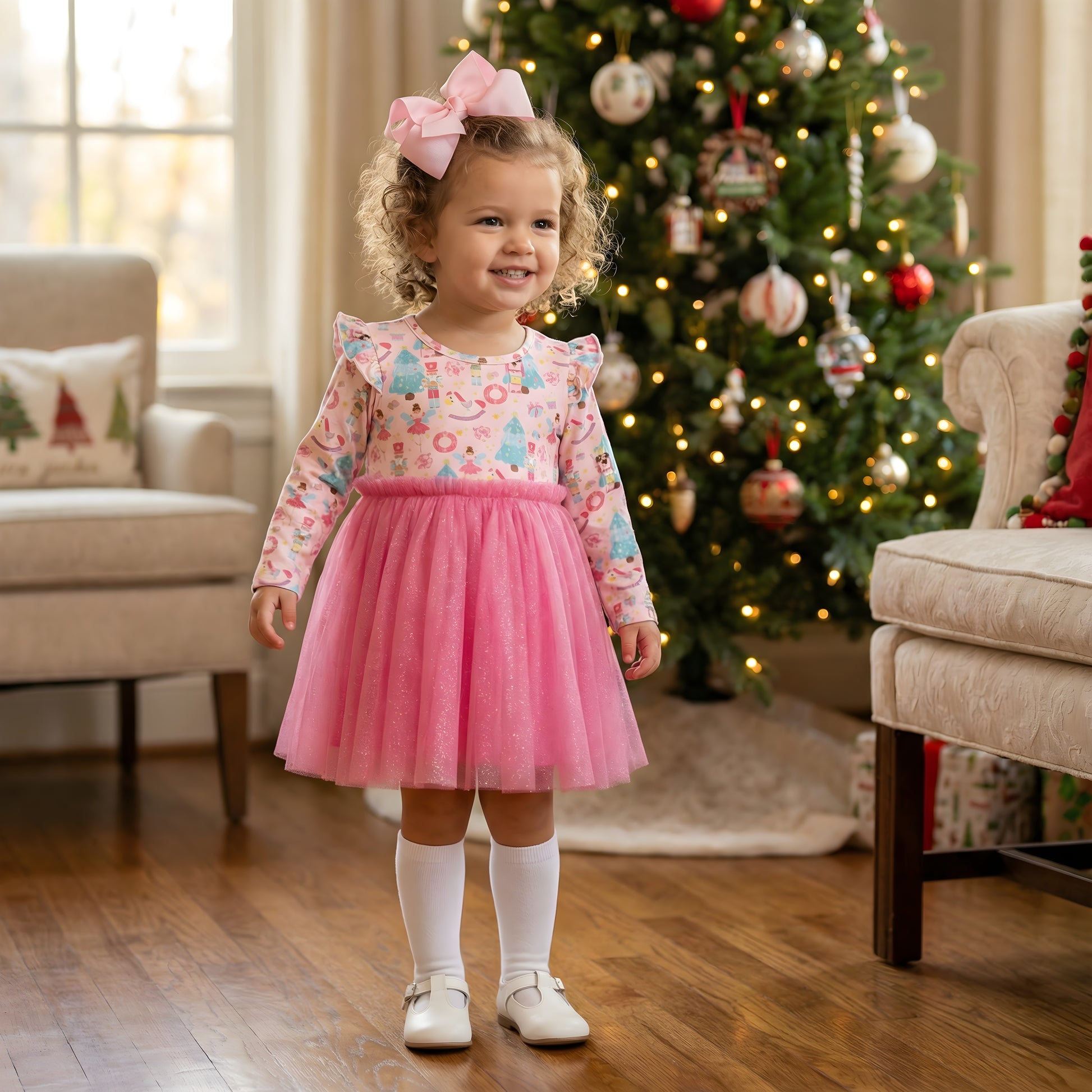 Toddler girl wearing a pink holiday dress with a tulle skirt and a long-sleeved bodice featuring nutcracker, candy, and Christmas tree motifs, complete with a large pink hair bow, white knee-high socks, and white Mary Jane shoes. She is standing on hardwood floors in front of a decorated Christmas tree.