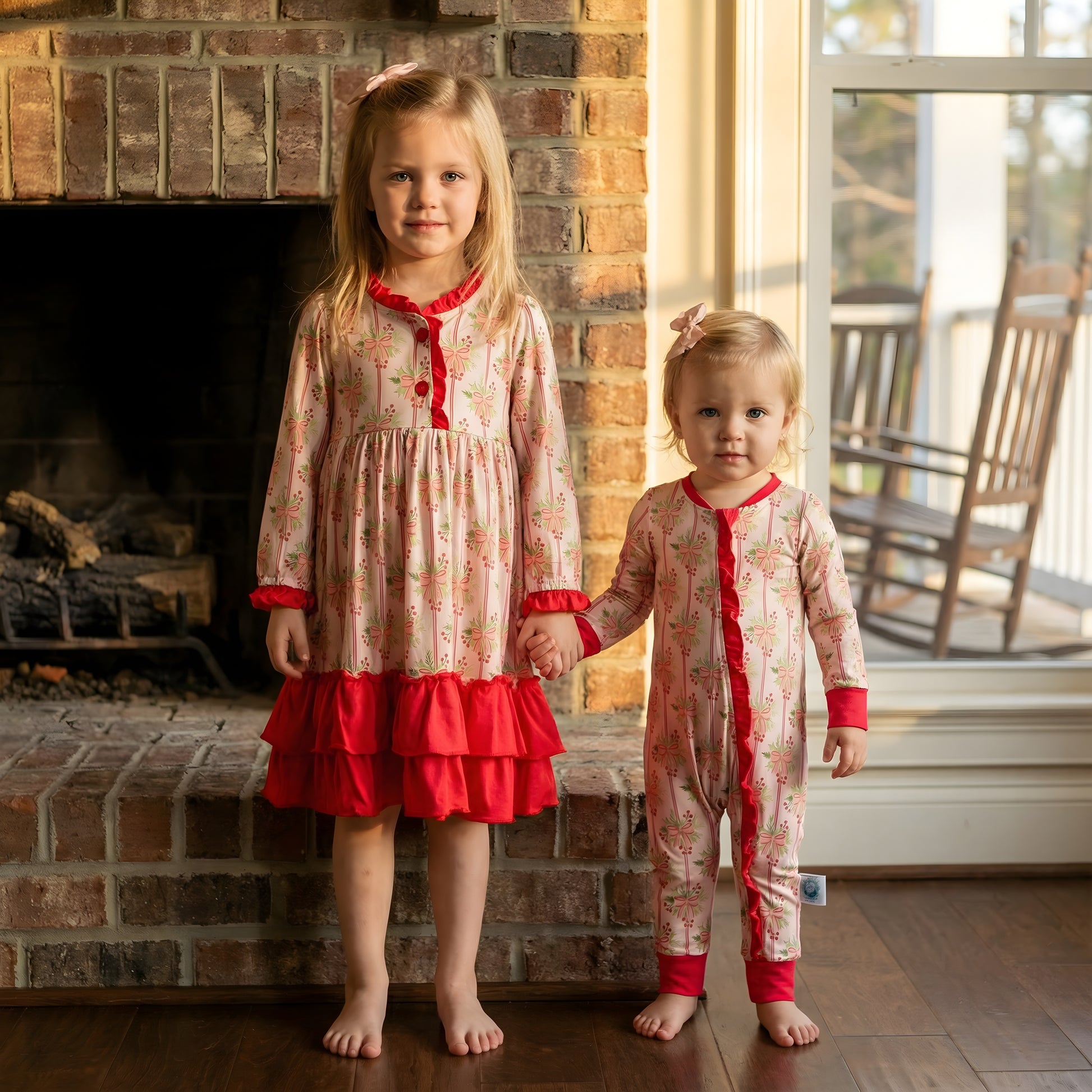 Two young girls, siblings matching in coordinating Bamboo pink Christmas sleepwear, stand hand-in-hand in front of a brick fireplace. The older girl wears a long-sleeve nightgown with red ruffles, while the younger wears a matching zipper onesie, both featuring a festive vintage ribbon and bow print.