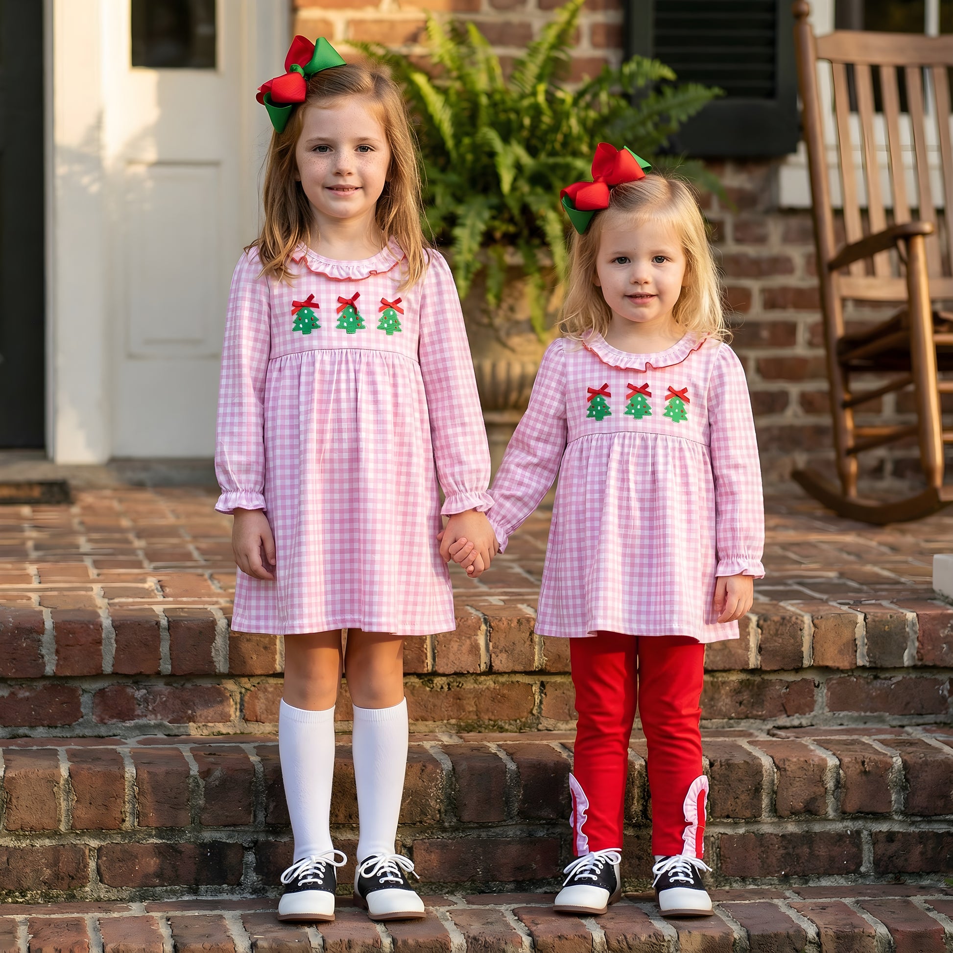 Two young girls standing on brick steps, wearing matching pink gingham smocked dresses with embroidered Christmas trees and red and green hair bows. The older girl wears white knee socks and saddle shoes, while the younger girl wears coordinating red ruffled leggings and saddle shoes. These outfits are a popular choice for holiday photos and Christmas clothing for sisters and toddlers.