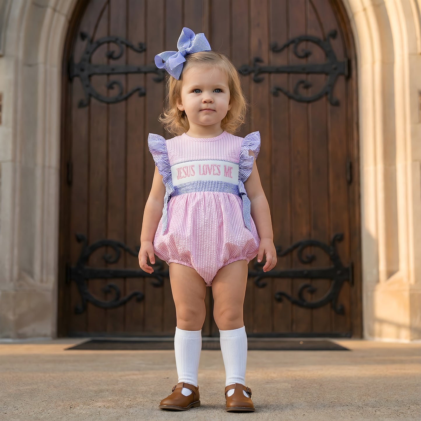 Toddler girl wearing pink seersucker Hand Smocked Girl Bubble - Jesus Loves Me with blue ruffled sleeves and matching hair bow standing in front of wooden church doors.