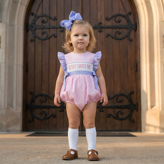 Toddler girl wearing pink seersucker Hand Smocked Girl Bubble - Jesus Loves Me with blue ruffled sleeves and matching hair bow standing in front of wooden church doors.