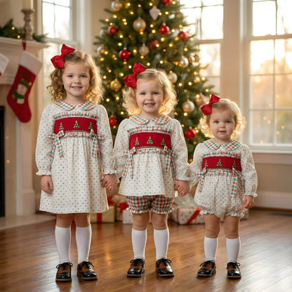 Three matching sibling girls pose in front of a decorated Christmas tree, wearing coordinating white Swiss Dot dresses and a romper with red hand smocked panels featuring Christmas tree designs.