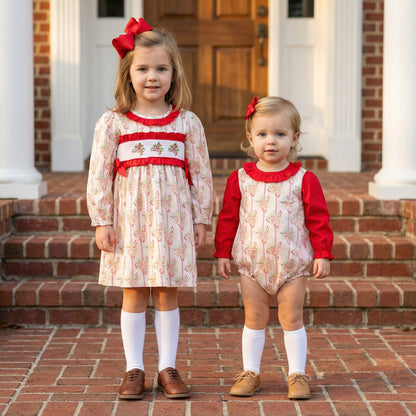 Two young girls stand on brick steps wearing coordinating holiday outfits in a cream and pink floral print with red accents. The older girl wears a long-sleeved smocked dress with a red ruffled collar and hair bow, while the younger girl wears a matching bubble romper with solid red sleeves. Both wear white knee-high socks and lace-up shoes.