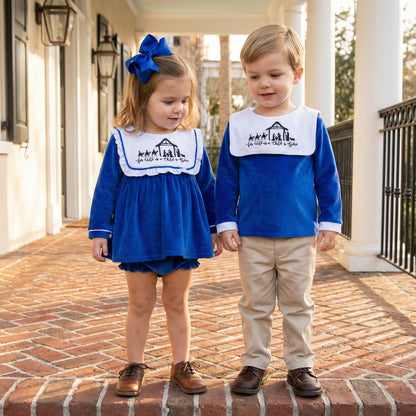 Two children standing on a brick walkway wearing matching royal blue holiday outfits with white bib-style collars embroidered with a nativity scene and the phrase ‘For Unto Us a Child is Born.’ The child on the left wears a long-sleeve blue top with matching diaper cover and a large blue hair bow, paired with brown shoes. The child on the right wears a long-sleeve blue top with khaki pants and brown shoes.