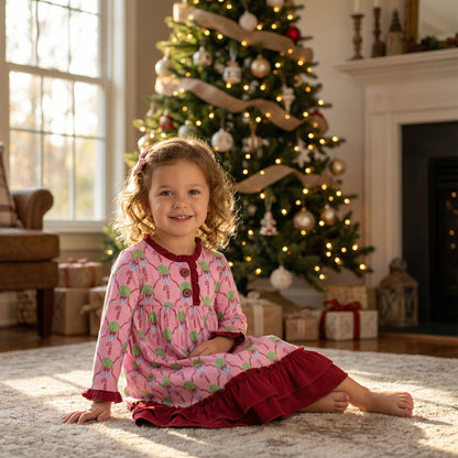 A young girl with curly light brown hair sits smiling on a plush beige rug, wearing a pink long-sleeved holiday nightgown featuring a candy cane pattern and deep red ruffled trim. Behind her is a cozy living room scene with a Christmas tree decorated with warm lights and burlap ribbon, wrapped gifts on the floor, and sunlight streaming through a large window.