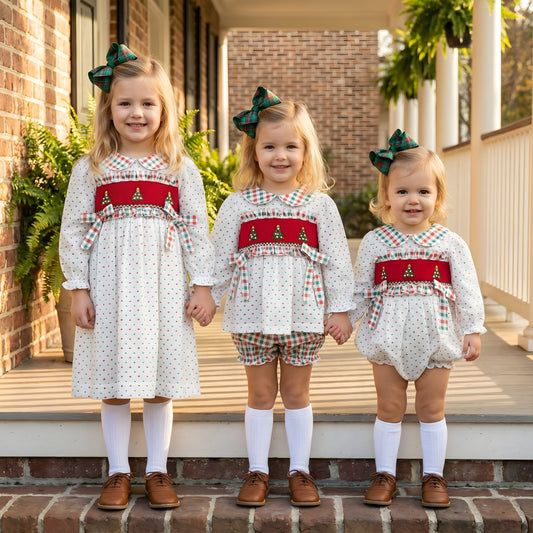 Three young girls wearing matching hand smocked Christmas outfits in white Swiss Dot fabric, featuring red smocking with tree embroidery and plaid bows.