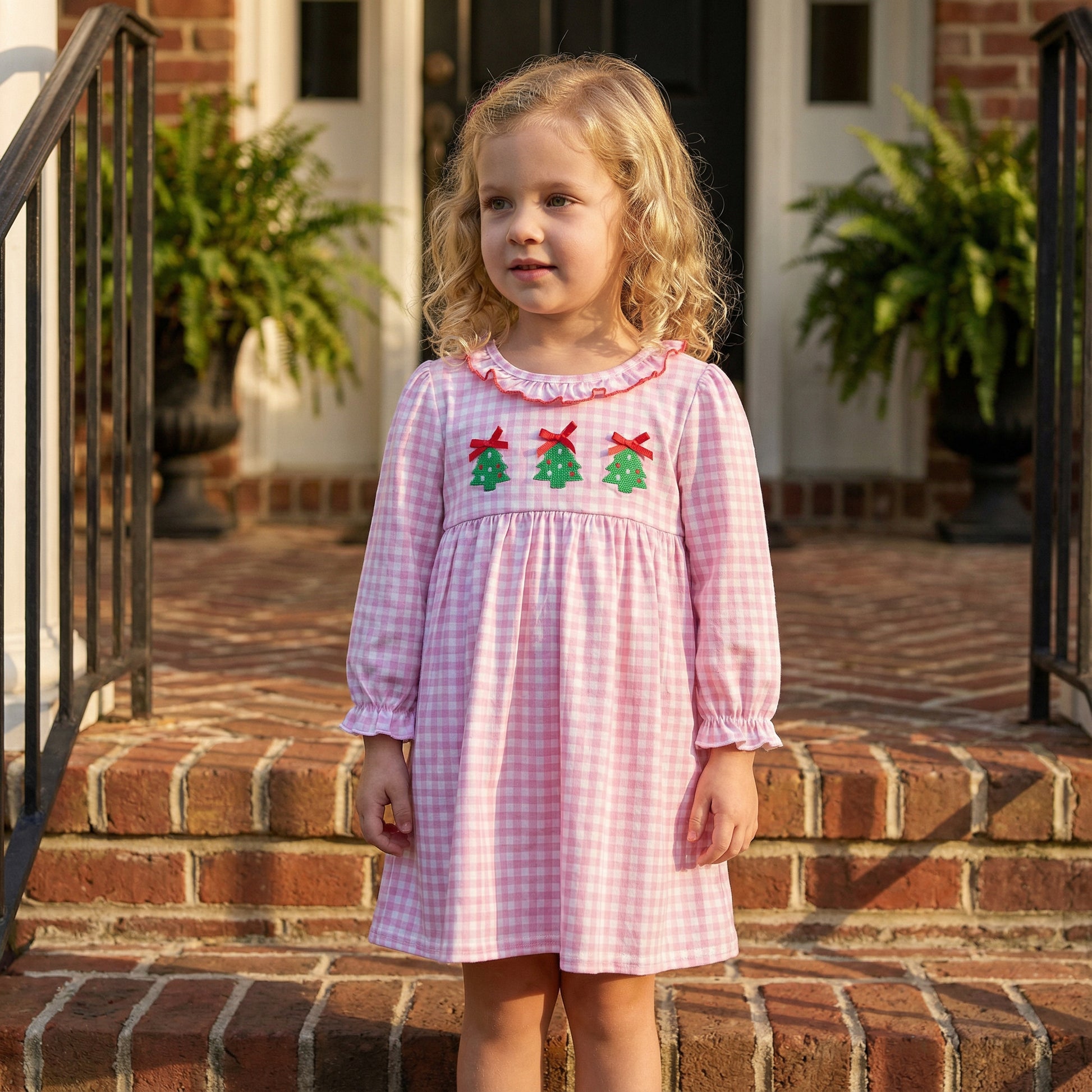 A young girl is wearing a pink and white gingham check dress with long sleeves and a ruffled collar. The bodice features smocking detailed with three embroidered green Christmas trees topped with red bows.